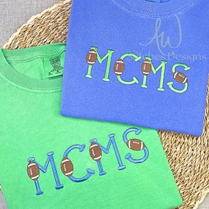 May include: Two green and blue t-shirts with the text "MCMS" embroidered in blue with a football graphic next to each letter.