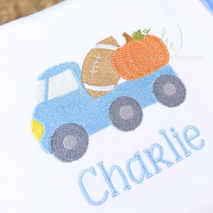 May include: Embroidered design of a light blue truck carrying a football and an orange pumpkin. The truck has gray wheels and a yellow headlight. The name "Charlie" is embroidered in blue script below the truck.
