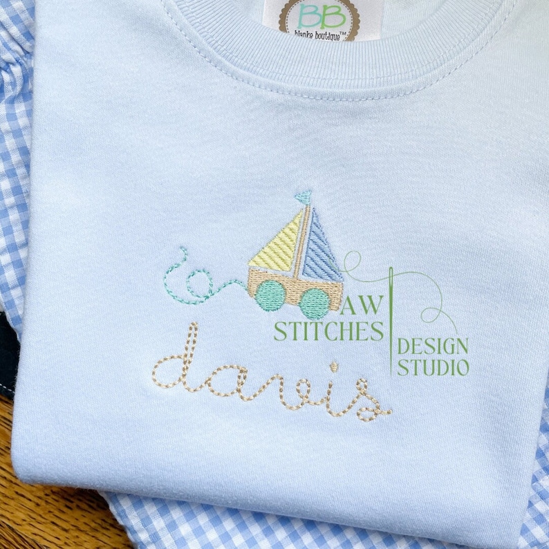 Pu&ograve; includere: Un body per neonato di colore azzurro chiaro con una barca a vela gialla e blu ricamata sul davanti. Il testo "AW STITCHES DESIGN STUDIO" e il nome "davis" sono anche ricamati sul body.