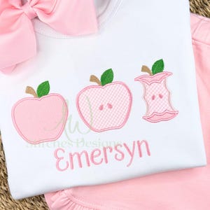 May include: White t-shirt with pink embroidered apples and the name "Emersyn" in pink script. The apples have green leaves and brown stems. A pink bow is at the top left, and pink shorts are visible on the right.
