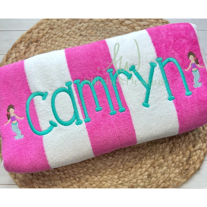 May include: A pink and white striped beach towel with the name "Camryn" embroidered in turquoise thread. Two small turquoise mermaids are embroidered on either side of the name.