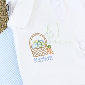 May include: A white embroidered shirt with a brown basket filled with colorful Easter eggs and carrots. The name "Nathan" is embroidered below the basket.