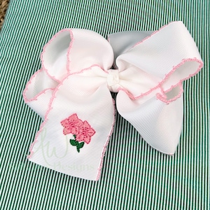May include: White hair bow with a pink flower embroidered on it. The bow has a pink scalloped edge.