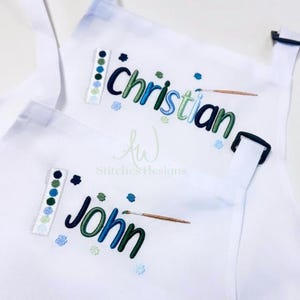 May include: White aprons embroidered with the names "Christian" and "John" in blue and green script. Each apron features a paint palette design and a paintbrush. The aprons are designed for artists or those who enjoy painting.