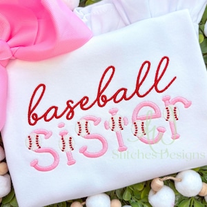 May include: White t-shirt with pink and red embroidery that says "baseball sister" with baseballs in the letters.