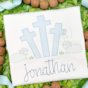 May include: White embroidered shirt with three blue crosses, a white lamb, and green grass. The name "Jonathan" is embroidered below the design.