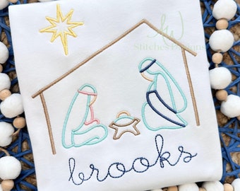 May include: A white embroidered Christmas ornament with a nativity scene and the name "brooks" in blue thread. The scene includes a star, a manger, Mary, Joseph, and baby Jesus.