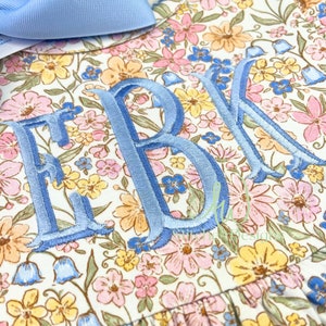Peut inclure: Une robe blanche &agrave; motif floral dans des tons de rose, jaune et bleu. La robe a un monogramme brod&eacute; bleu "EBK" sur le devant.