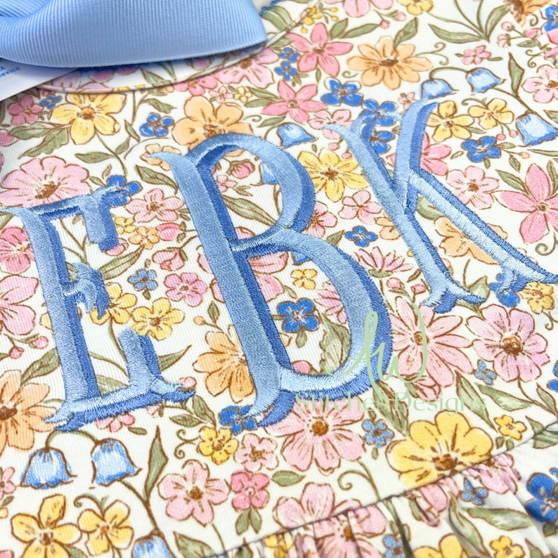 Peut inclure: Une robe blanche &agrave; motif floral dans des tons de rose, jaune et bleu. La robe a un monogramme brod&eacute; bleu "EBK" sur le devant.