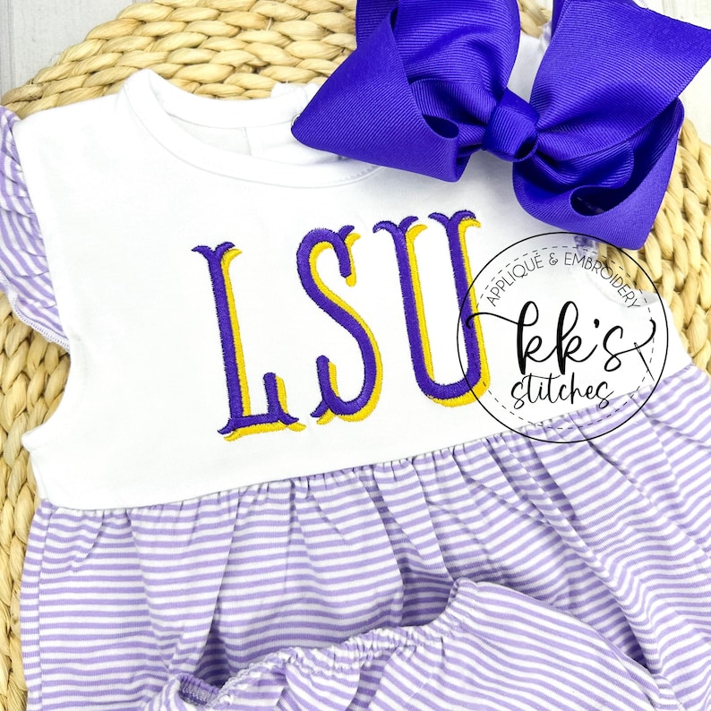 Peut inclure: Une robe de b&eacute;b&eacute; ray&eacute;e blanche et violette avec un corsage blanc orn&eacute; des lettres brod&eacute;es "LSU" en violet et jaune. La robe a un col &agrave; volants et un n&oelig;ud en satin violet.