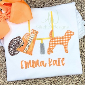 May include: White t-shirt with orange football, goalpost, and dog applique. The word "Tennessee" is on a pennant. The name "Emma Kate" is embroidered in orange. An orange bow is in the upper left corner.