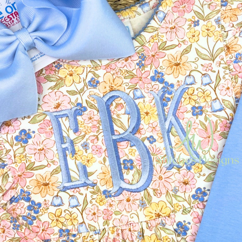 Peut inclure: Une robe blanche &agrave; imprim&eacute; floral dans des tons de rose, jaune et bleu. La robe a un col rond et un monogramme des lettres "EBK" brod&eacute; sur le devant en fil bleu clair.