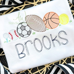 May include: White embroidered t-shirt with a design of various sports balls, including a football, basketball, soccer ball, baseball, and tennis ball. The name "Brooks" is embroidered below the balls.