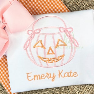 Puede incluir: Un pelele blanco con una calabaza bordada en rosa y naranja con una cara de calabaza y el nombre "Emery Kate" bordado debajo.