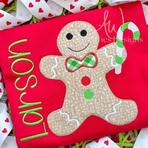 May include: Red t-shirt with a gingerbread man applique holding a candy cane. The gingerbread man has green and red accents and the text "Fair is fun" is embroidered in green on the shirt.