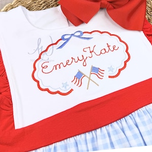 May include: White and red patriotic outfit with a large red bow. The outfit features a white top with the name "Emery Kate" embroidered in red, along with blue flags and stars. The bottom has a blue and white gingham pattern.