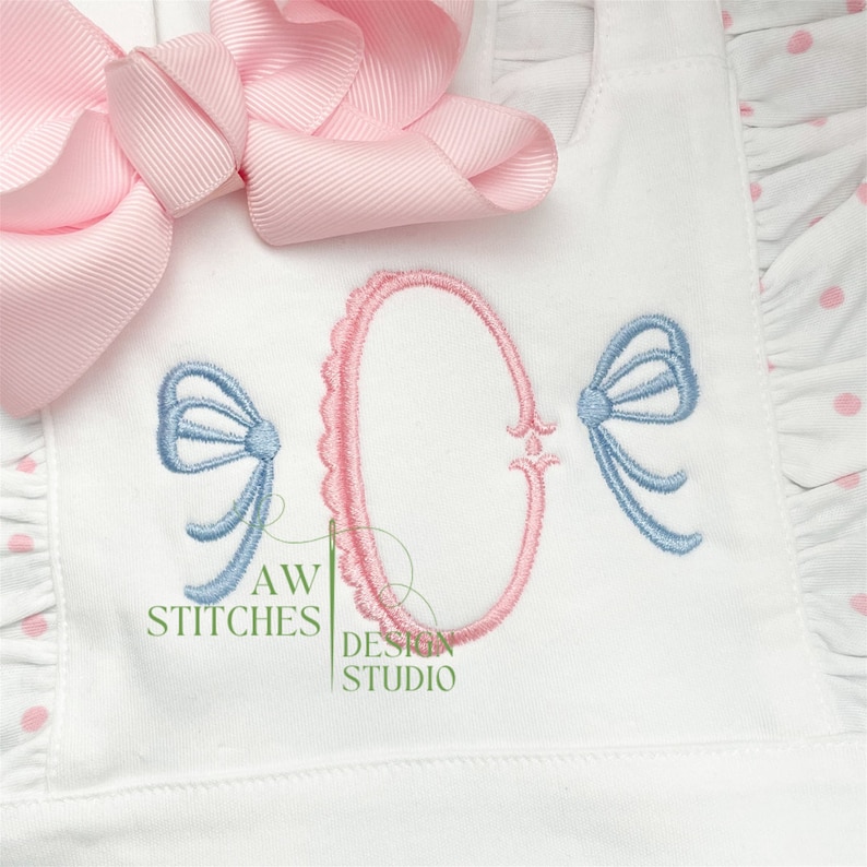 May include: A white fabric with a pink and blue embroidered design. The design features a large oval with a scalloped edge, two smaller bows, and the text "AW STITCHES DESIGN STUDIO".
