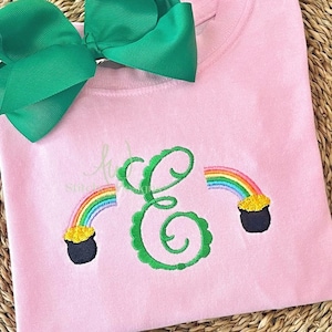 May include: Pink sweatshirt with a large green bow at the top. The sweatshirt features a green embroidered letter "E" with two rainbows and pots of gold on either side. The rainbows are multi-colored.