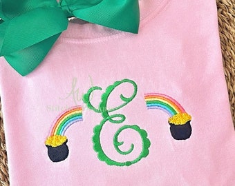 May include: Pink sweatshirt with a large green bow at the top. The sweatshirt features a green embroidered letter "E" with two rainbows and pots of gold on either side. The rainbows are multi-colored.