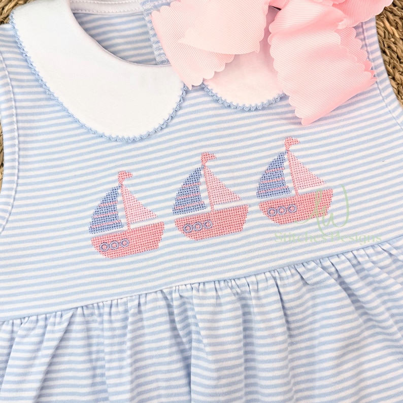 Op de afbeelding: Een mouwloze babyjurk met blauw-witte strepen, met een borduursel van drie roze en blauwe zeilboten. De jurk heeft een witte kraag en een grote roze strik. De stof is zacht en licht.