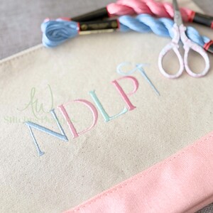 May include: A cream-colored canvas tote bag with the embroidered letters "NDLP" in pastel blue and pink. The bag has a pink trim and is accompanied by blue and pink embroidery floss and small scissors.