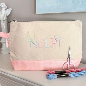 May include: A canvas cosmetic bag with a pink bottom and a zipper closure. The bag features embroidered initials in pastel colors and a needle and thread design. A pair of small scissors and two spools of thread are next to the bag.