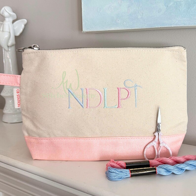 May include: A canvas cosmetic bag with a pink bottom and a zipper closure. The bag features embroidered initials in pastel colors and a needle and thread design. A pair of small scissors and two spools of thread are next to the bag.