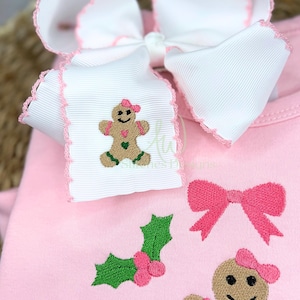 Op de afbeelding: Een lichtroze onesie met geborduurde kerstthema-ontwerpen. Het ontwerp omvat een peperkoeken mannetje, een roze strik en hulst. Een grote witte strik met roze rand is aan de onesie bevestigd.