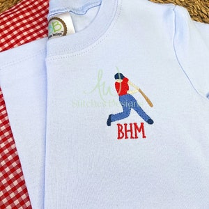 May include: A light blue t-shirt with a red, white, and blue embroidered baseball player design. The player is swinging a bat and the name "BHM" is embroidered below the player.