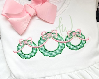 Op de afbeelding: Witte, mouwloze jurk met drie groene en witte gingham-kransen met roze strikken. De kransen zijn verbonden door een roze lint.