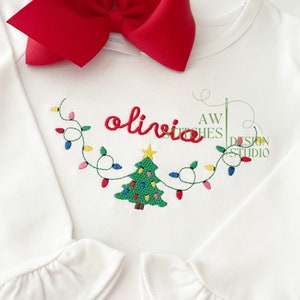 May include: White long-sleeve shirt with a red bow on the top. The shirt has an embroidered design of a Christmas tree with lights and the name "olivia" above it.
