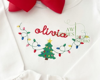 May include: White long-sleeve shirt with a red bow on the top. The shirt has an embroidered design of a Christmas tree with lights and the name "olivia" above it.