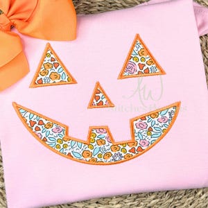 May include: Pink Halloween-themed shirt with an embroidered jack-o'-lantern face. The eyes and mouth are filled with a floral pattern in orange, pink, and blue. An orange bow is visible in the upper left corner. The text "AW Stitches Plus" is visible.