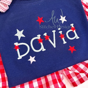 May include: A navy blue t-shirt with the name "DAVID" embroidered in white with red stars around the name and scattered on the shirt. The shirt is folded over a red and white gingham fabric.
