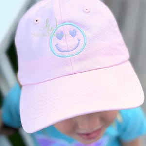May include: A light pink baseball cap with a curved brim. The cap features an embroidered smiley face with heart-shaped eyes in light blue. The text "Stitches" is visible on the side of the cap.