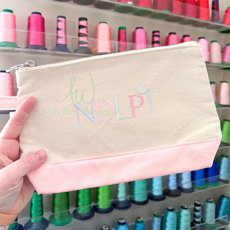 May include: A beige canvas zippered pouch with a light pink bottom panel and a matching handle. The pouch features embroidered initials in pastel colors. The background shows spools of thread in various colors.