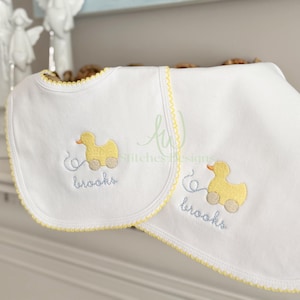 May include: Two white baby bibs with yellow trim and a yellow duck design. The duck has wheels and is embroidered with the name "brooks" in light blue. The bibs have a scalloped edge.