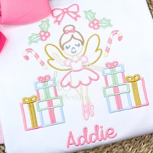 May include: White t-shirt embroidered with a ballerina, gifts, candy canes, and holly. The ballerina is wearing a pink tutu and ballet shoes. The name "Addie" is embroidered in pink script below the design. A large pink bow is visible.