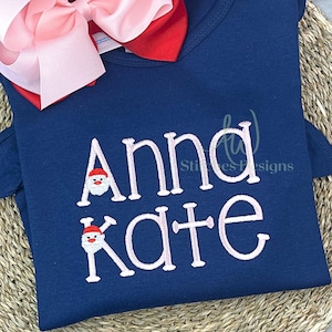 May include: A navy blue shirt with the name "Anna Kate" embroidered in pink with a Santa Claus face in each of the letters "A" and "K".