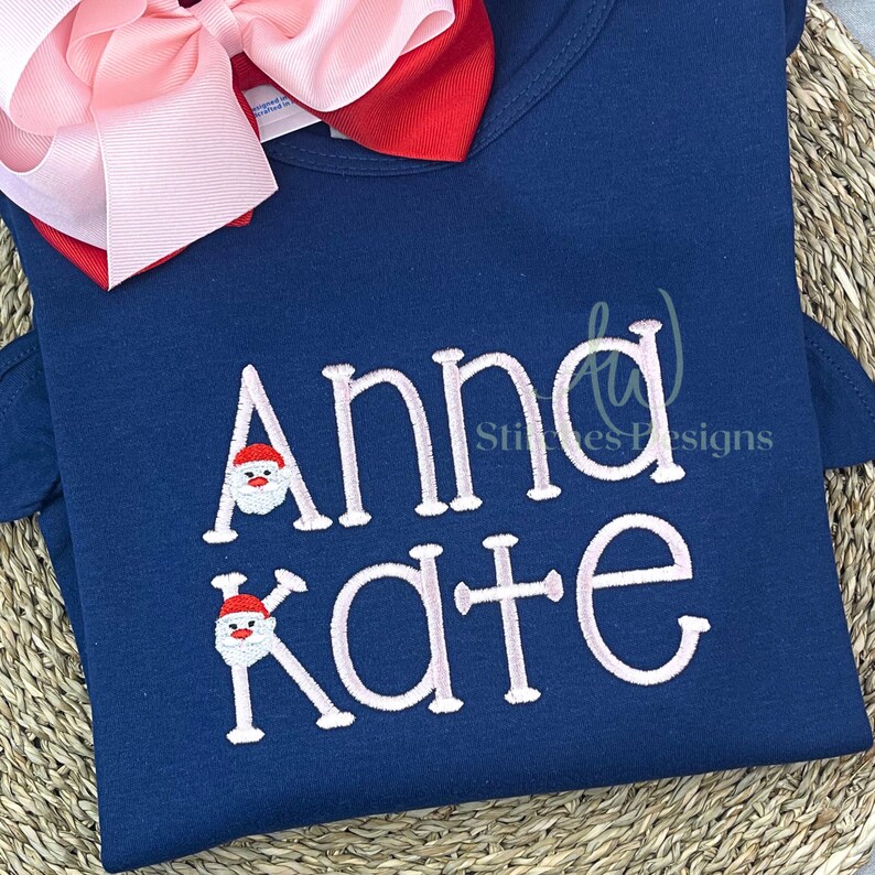May include: A navy blue shirt with the name "Anna Kate" embroidered in pink with a Santa Claus face in each of the letters "A" and "K".