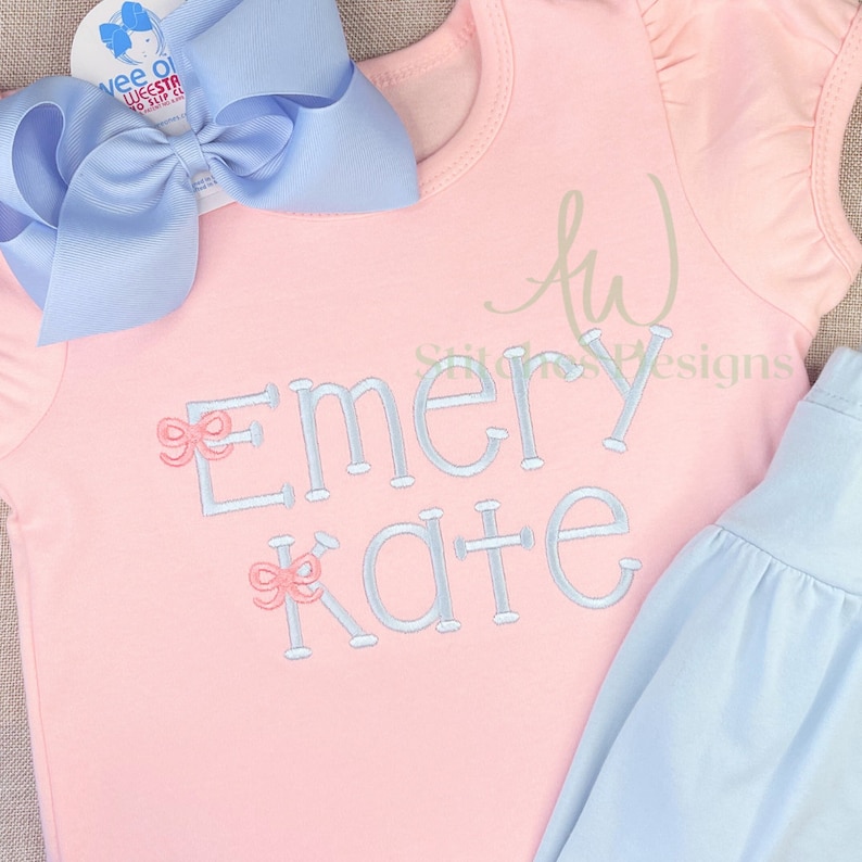 May include: A pink short-sleeved shirt with the name "Emery Kate" embroidered in light blue with pink bows. The shirt is paired with a light blue skirt.