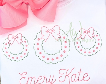 Op de afbeelding: Wit T-shirt met drie groene geborduurde kransen met roze strikken. De naam "Emery Kate" is in roze onder de kransen geborduurd.