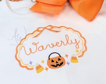 May include: A white short-sleeved bodysuit featuring an orange embroidered scalloped frame with the name Waverly in cursive script. Below the name is an embroidered jack-o'-lantern bucket, candy corn, and small stars. A large orange hair bow is attached.