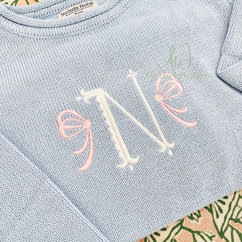 May include: A light blue knitted sweater with a white embroidered letter "N" and pink bows on either side of the letter.