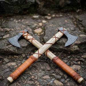 May include: Two crossed axes with wooden handles wrapped in brown leather. The axe heads have a patterned design. The axes are resting on a stone surface.
