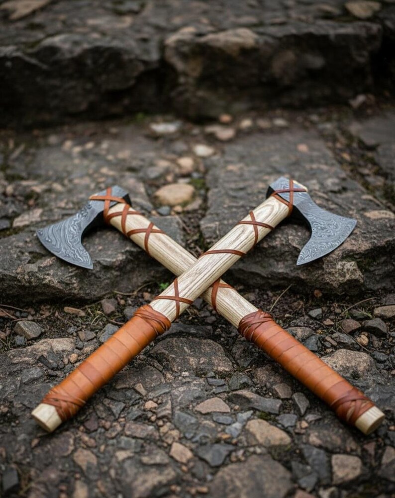 May include: Two crossed axes with wooden handles wrapped in brown leather. The axe heads have a patterned design. The axes are resting on a stone surface.