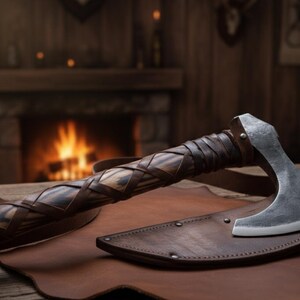 May include: A handcrafted axe with a leather-wrapped handle and a steel blade, resting on a leather sheath. The handle features a woven design. The axe is displayed on a wooden surface, suggesting a rustic or historical theme.