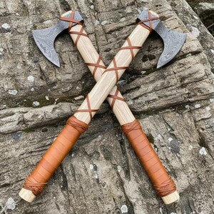 May include: Two crossed axes with light wood handles and patterned steel blades. The handles are wrapped with brown leather, and the axes are resting on a textured rock surface. The axes have a rustic, handcrafted appearance.