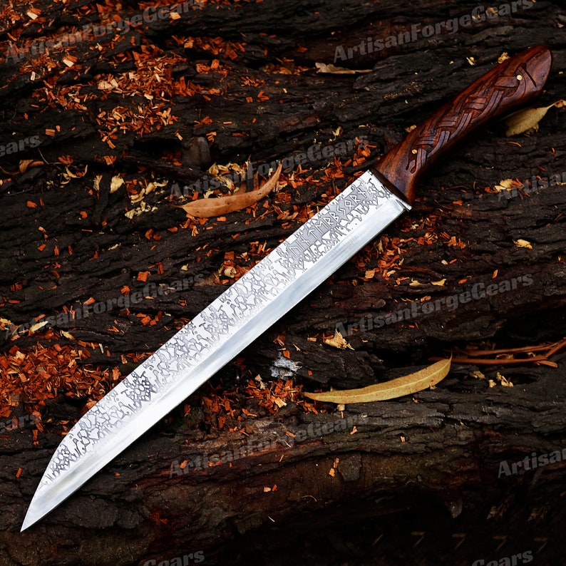 May include: A long, silver knife with a wooden handle. The handle is carved with a knot design. The blade is etched with a pattern. The knife is lying on a wooden surface covered in wood chips.