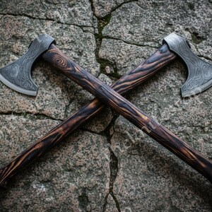 May include: Two crossed axes with intricately carved steel blades and dark wooden handles. The handles have a unique, burnt wood grain pattern. The axes are displayed on a textured, gray stone surface.